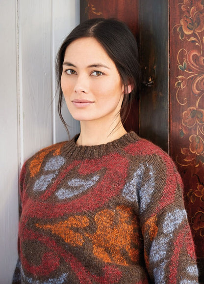 A woman with dark hair and an orange, brown, and blue patterned sweater leans against a wooden wall with ornate floral designs, offering a soft smile.