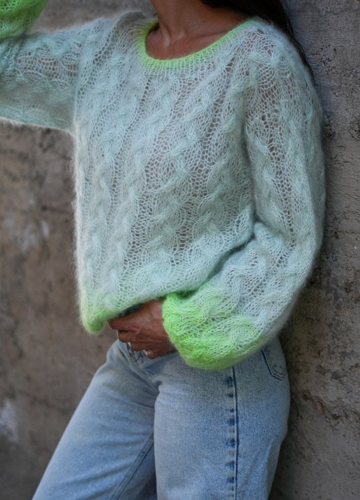 A person wearing a fluffy, pastel green knit sweater with neon green accents stands against a textured wall, paired with light blue jeans. Cozy and stylish mood.