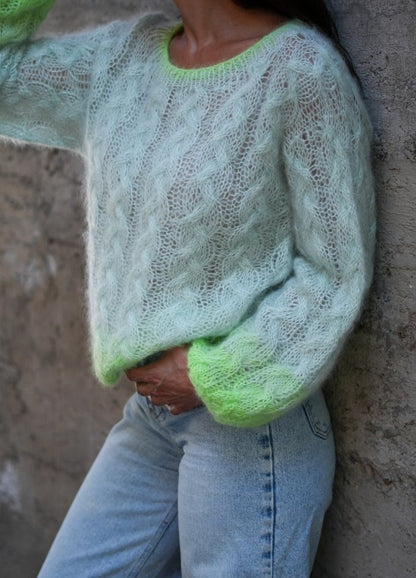 A person wearing a fluffy, pastel green knit sweater with neon green accents stands against a textured wall, paired with light blue jeans. Cozy and stylish mood.