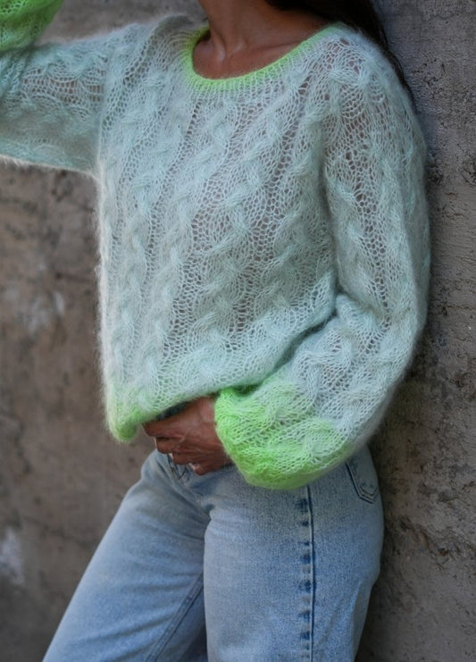 A person wearing a fluffy, pastel green knit sweater with neon green accents stands against a textured wall, paired with light blue jeans. Cozy and stylish mood.