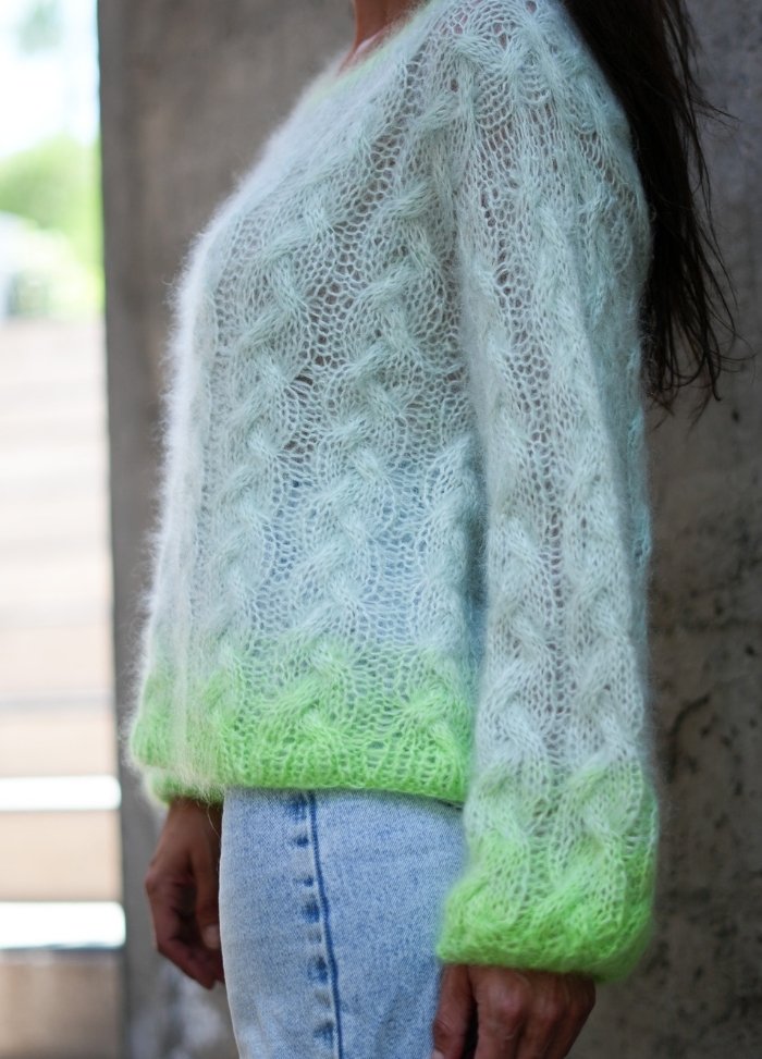 A person wearing a light blue and neon green ombre knit sweater stands sideways against a concrete wall, creating a cozy and stylish vibe.