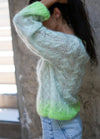 A person wearing a soft, translucent blue sweater with green gradient detail at the hem, paired with blue jeans. The scene is calm and casual.