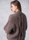 A woman with long brown hair is turned to the side, wearing a thick, textured brown sweater with intricate braided cable patterns. The background is soft gray.