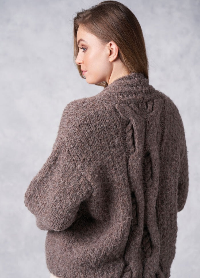 A woman with long brown hair is turned to the side, wearing a thick, textured brown sweater with intricate braided cable patterns. The background is soft gray.