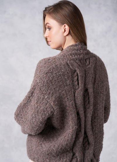 A woman with long brown hair is turned to the side, wearing a thick, textured brown sweater with intricate braided cable patterns. The background is soft gray.