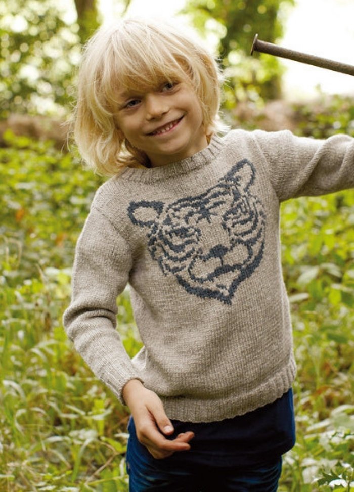 Child in light gray Tiger Sweater with tiger face motif, outdoors with plants.
