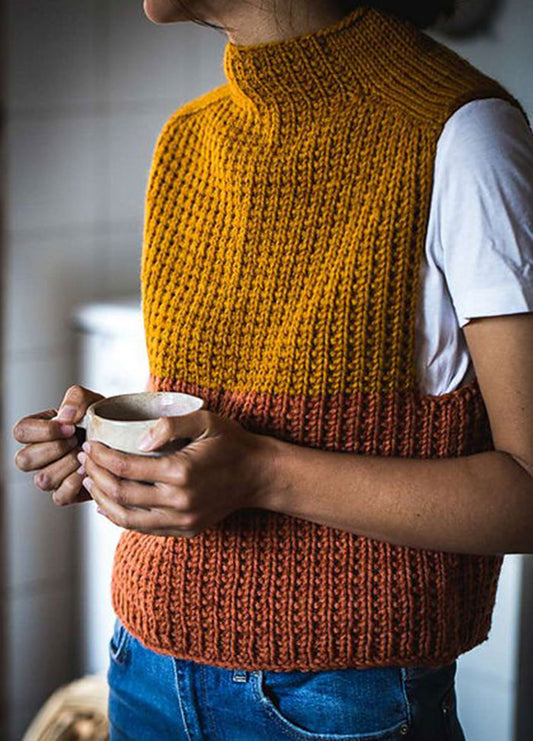 Timbo Vest featuring a ribbed knit texture with a high neck, color-blocked in mustard yellow and rust orange, worn over a white short-sleeved shirt with blue jeans. The model holds a ceramic cup, showcasing the vest’s cozy and casual styling.