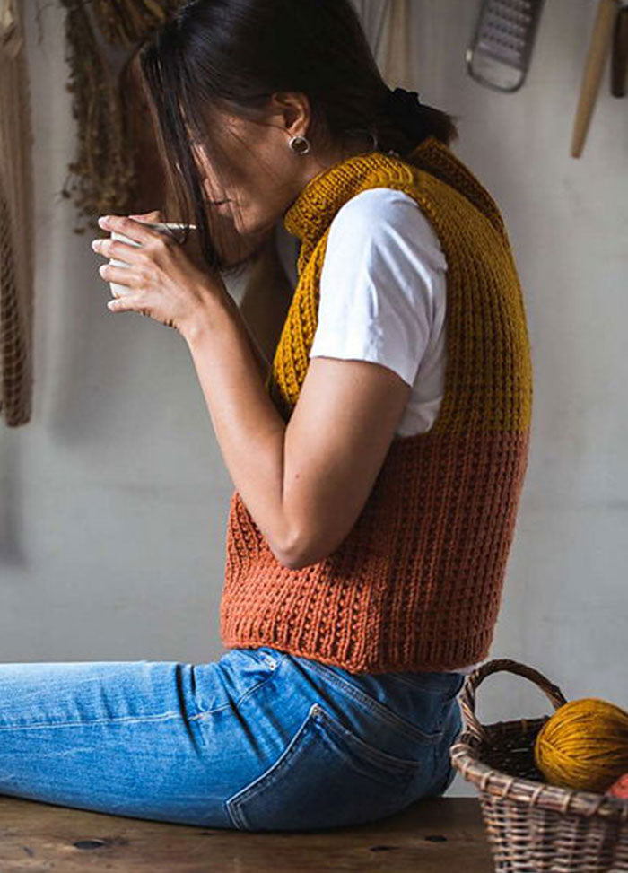 Timbo Vest in mustard yellow and burnt orange, hand-knitted with a ribbed texture, worn over a white short-sleeved shirt and blue jeans. The person is sitting on a wooden surface, holding a cup near their face, with a basket of yarn balls beside them.