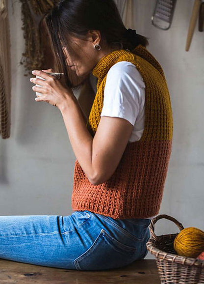 Timbo Vest in mustard yellow and burnt orange, hand-knitted with a ribbed texture, worn over a white short-sleeved shirt and blue jeans. The person is sitting on a wooden surface, holding a cup near their face, with a basket of yarn balls beside them.