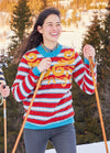 A person smiles while skiing in a vibrant red and blue striped sweater with yellow patterns. Snowy landscape and pine trees are in the background.