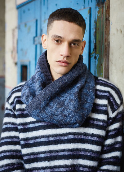 A person stands against a rustic blue door, wearing a chunky blue scarf and a striped sweater. The expression is calm, evoking a cool, urban vibe.
