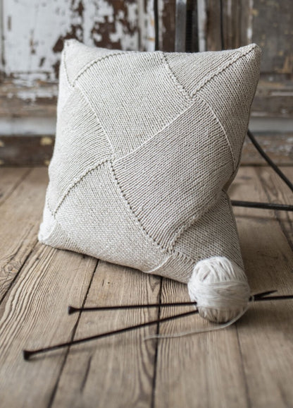 Cream-colored knit pillow with a geometric pattern resting on a rustic wooden floor. Nearby, a yarn ball and knitting needles convey a cozy, handcrafted vibe.