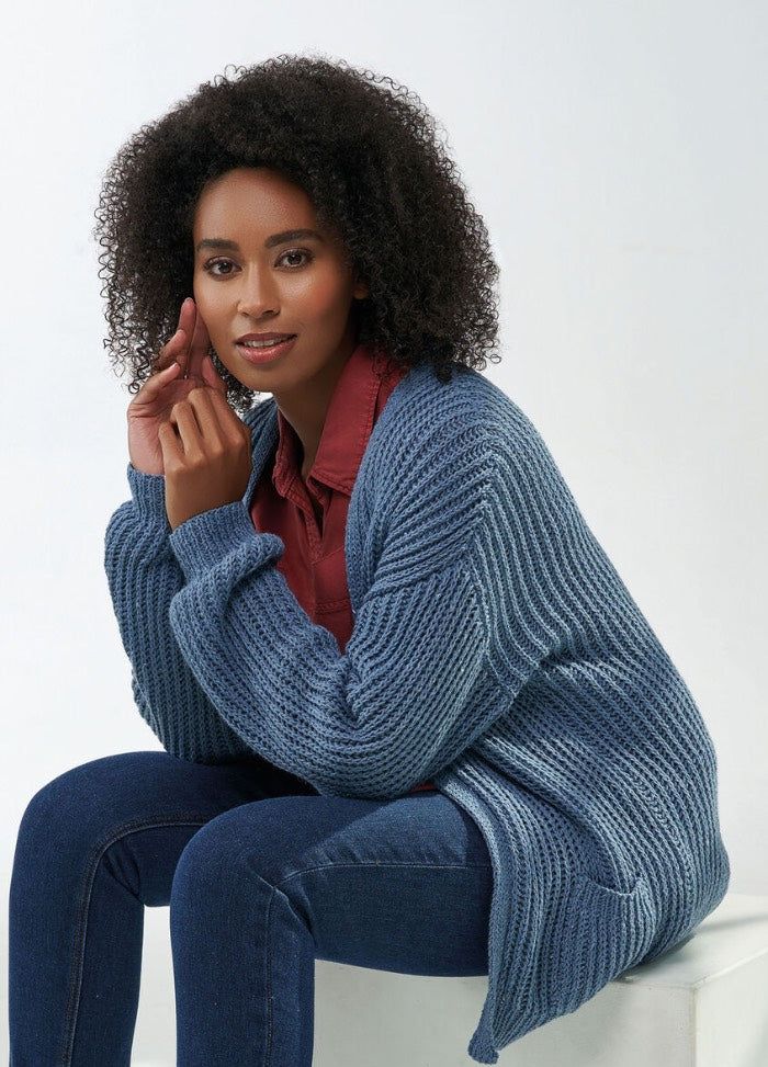 Woman with curly hair sits, wearing a red shirt and blue knit cardigan. She rests her chin on her hand, conveying a calm, thoughtful demeanor.