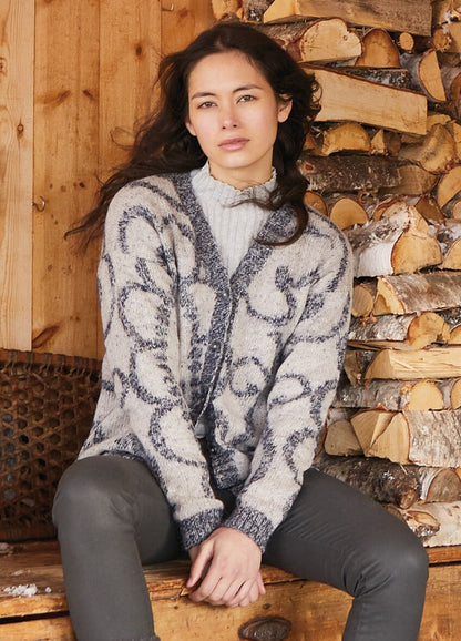 A woman in a cozy, patterned cardigan sits on a wooden bench beside stacked firewood. She has long brown hair and wears a neutral expression.