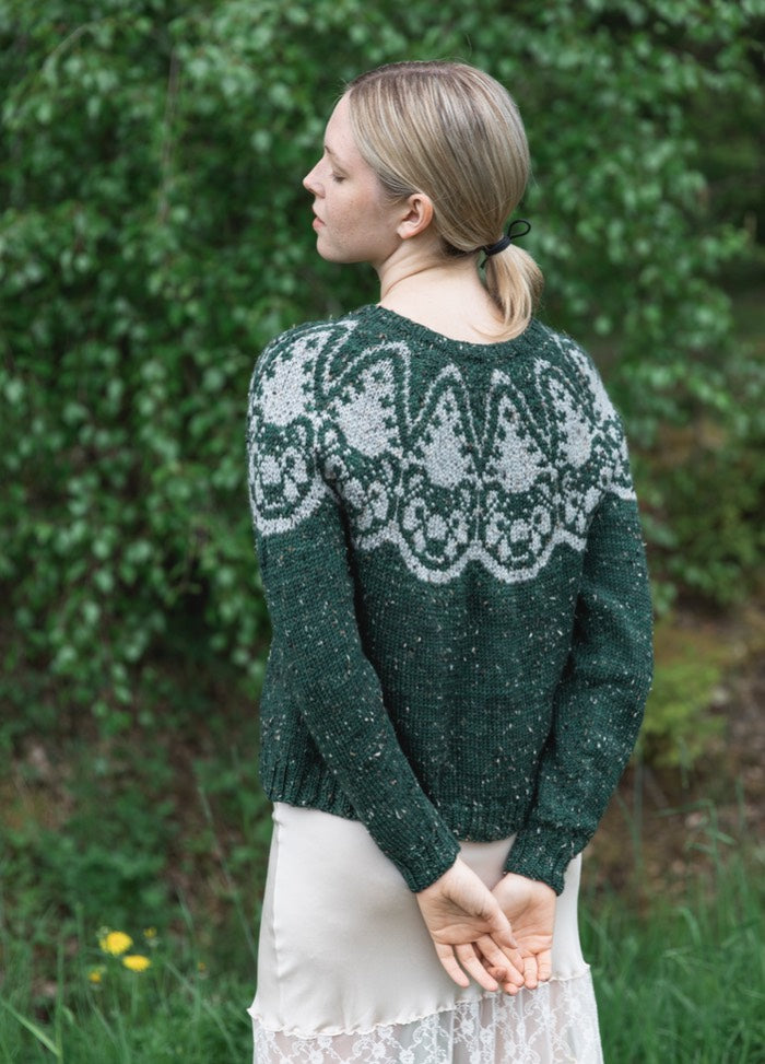 Tuonen Karhu Sweater: dark green knit with light grey motifs across the shoulders and upper back, shown from behind in a leafy outdoor setting.