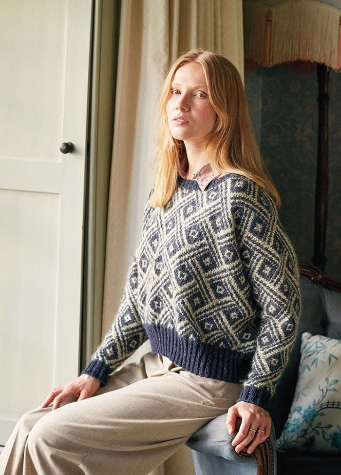 A woman with long blonde hair sits on a chair by a window, wearing a patterned blue and white sweater and beige pants, conveying a calm, cozy vibe.