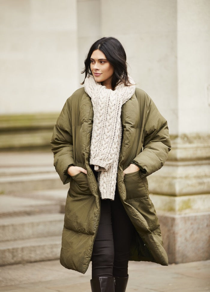A woman wearing a long, olive green puffer coat and a thick, cream-colored knit scarf stands with hands in pockets. She looks relaxed, exuding warmth and confidence.