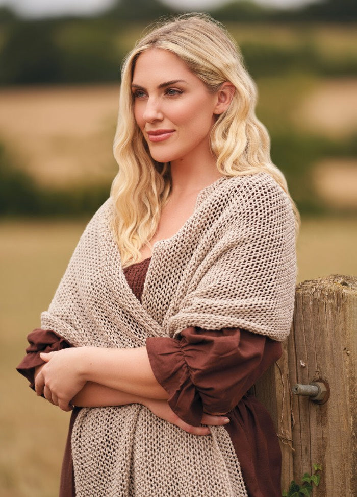 A woman with long blonde hair stands in a serene field, wearing a brown dress and beige shawl. She leans on a wooden post, looking thoughtful.