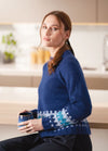 A person with brown hair sits in a modern kitchen, wearing a blue sweater with white and teal patterns. They hold a blue mug, exuding a calm demeanor.