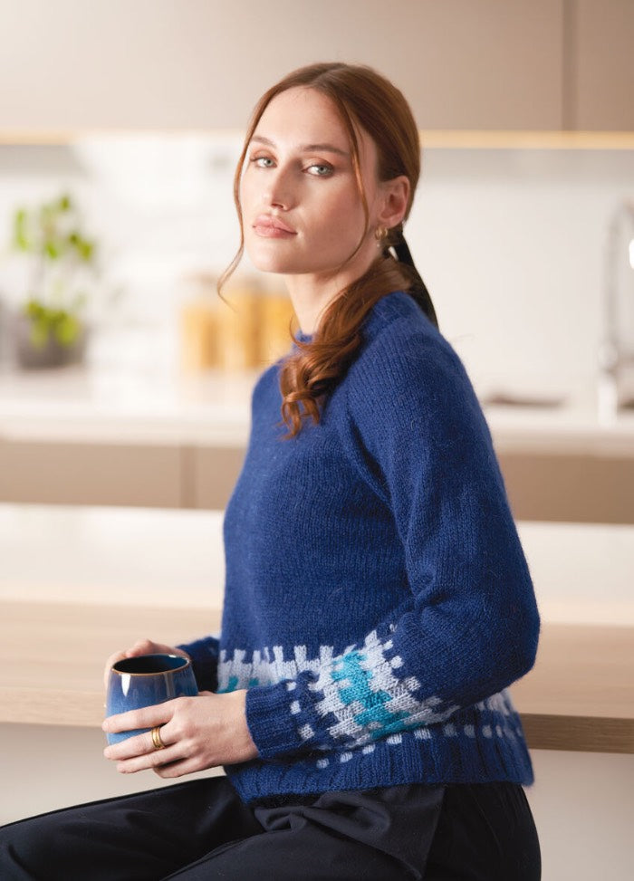 A person with brown hair sits in a modern kitchen, wearing a blue sweater with white and teal patterns. They hold a blue mug, exuding a calm demeanor.