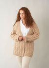 A woman with long curly auburn hair wears a chunky beige cardigan with a deep V-neckline, buttoned front, and textured cable pattern. She pairs it with a crisp white shirt and light trousers, standing against a plain neutral background.