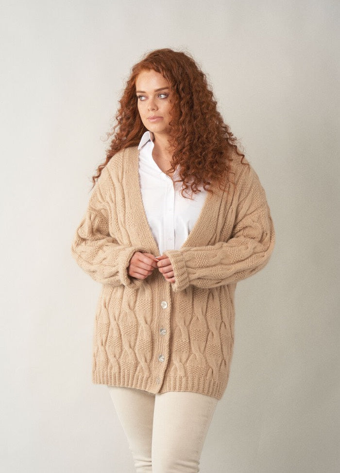 A woman with long curly auburn hair wears a chunky beige cardigan with a deep V-neckline, buttoned front, and textured cable pattern. She pairs it with a crisp white shirt and light trousers, standing against a plain neutral background.