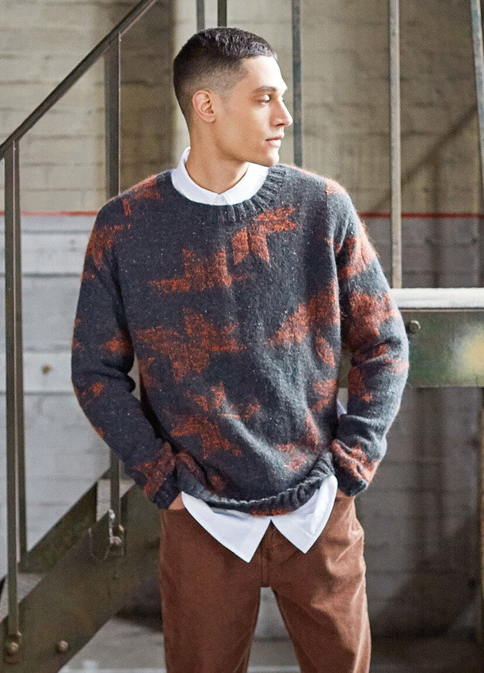 Young man wearing a gray and orange patterned sweater over a white shirt and brown pants, standing by staircase, gazing to the side, in a relaxed pose.