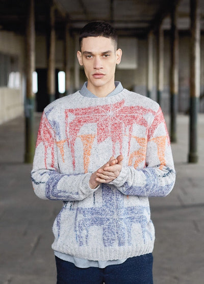 A man stands in an industrial setting, wearing a colorful patterned sweater. He has a serious expression and clasps his hands. The scene feels contemplative.