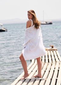 A woman in a white dress and cardigan stands barefoot on a wooden pier, gazing at the calm sea. The setting is serene with distant boats.