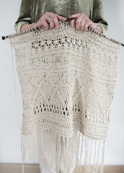 A person in a green sweater holds knitting needles with an intricate, cream-colored knitted piece featuring lace patterns and fringe hanging below.