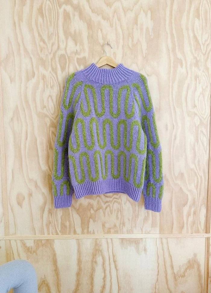 Purple sweater with green geometric patterns hanging on a wooden wall. The textured background adds a rustic feel to the cozy knit design.