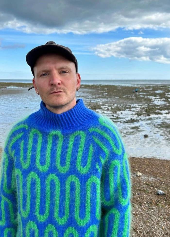 A person stands on a pebbly beach wearing a vibrant blue and green patterned sweater and a black cap, under a cloudy sky with patches of blue.