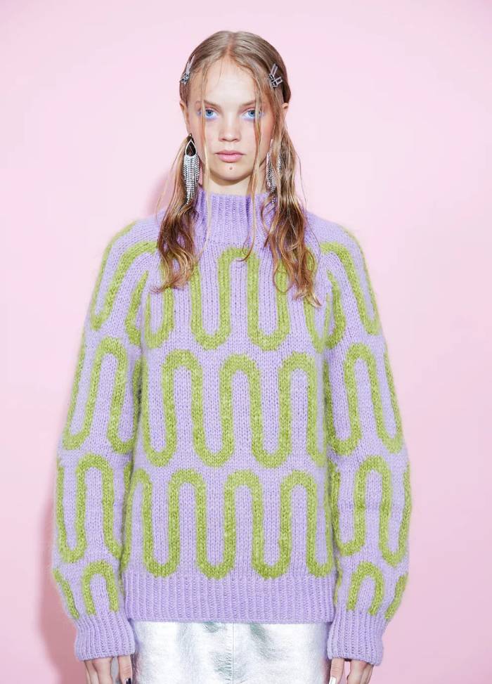 Young woman stands against a pink background, wearing a purple sweater with green patterns and metallic skirt. Her styled hair conveys a trendy, modern vibe.