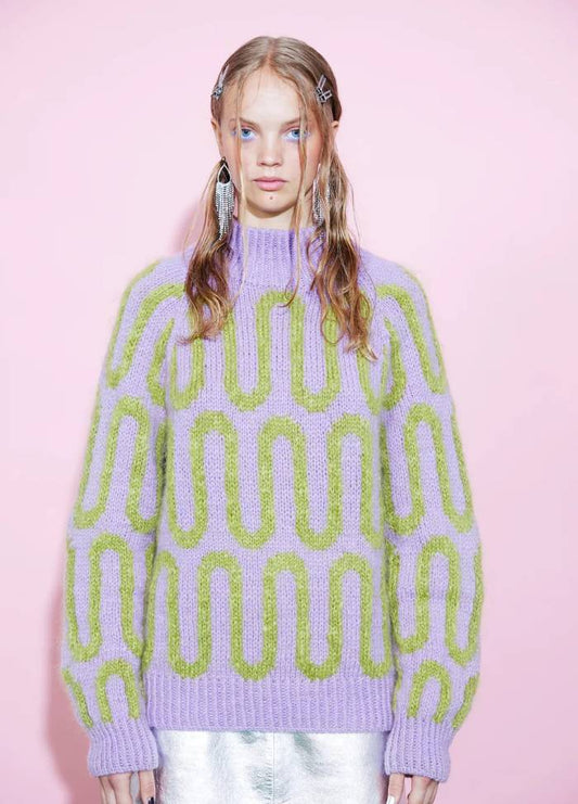 Young woman stands against a pink background, wearing a purple sweater with green patterns and metallic skirt. Her styled hair conveys a trendy, modern vibe.