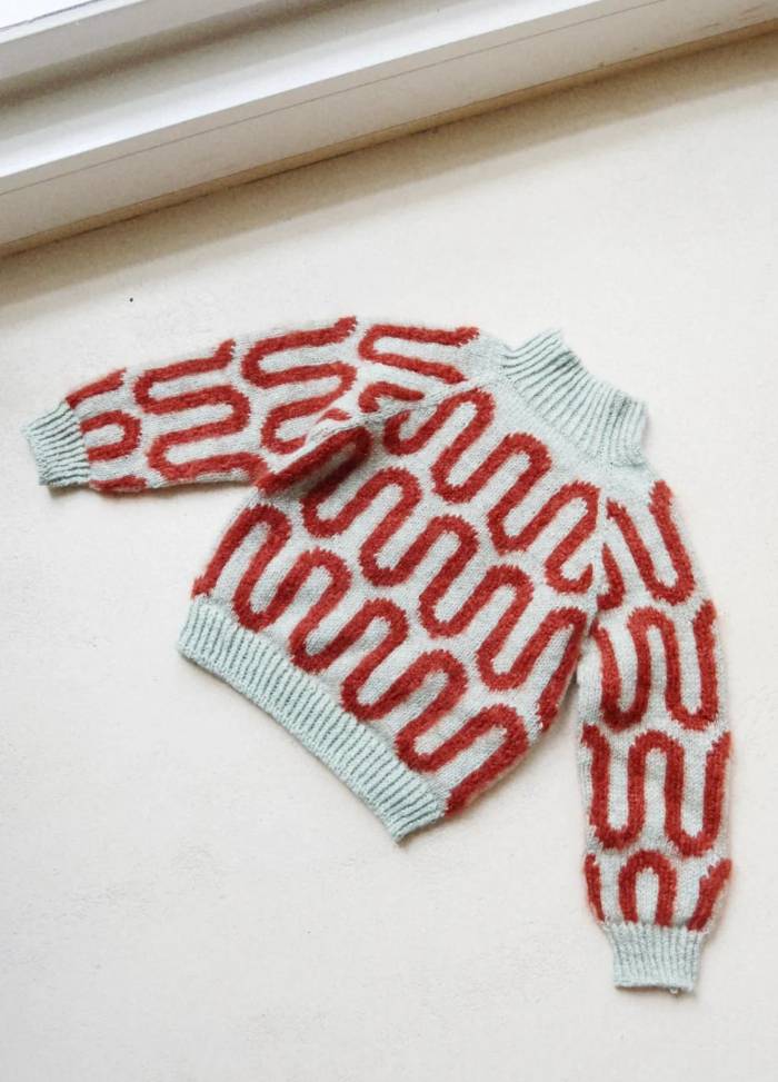 A cozy wool sweater lies flat on a light surface. It features a mint green base with bold, red loop patterns and ribbed cuffs and neckline.