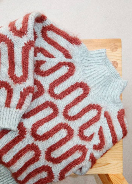 Cozy light blue knit sweater with red wavy patterns placed on a wooden chair. The texture appears soft, conveying warmth and comfort.