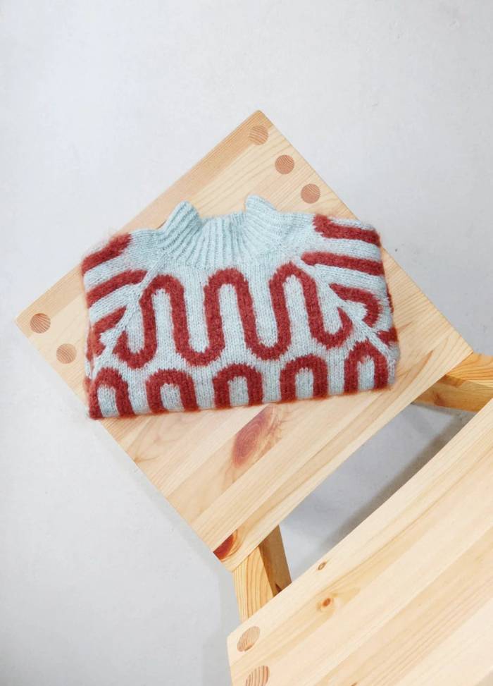 A folded sweater with a light blue base and red wavy patterns lies on a wooden chair. The setting is minimalistic and cozy.