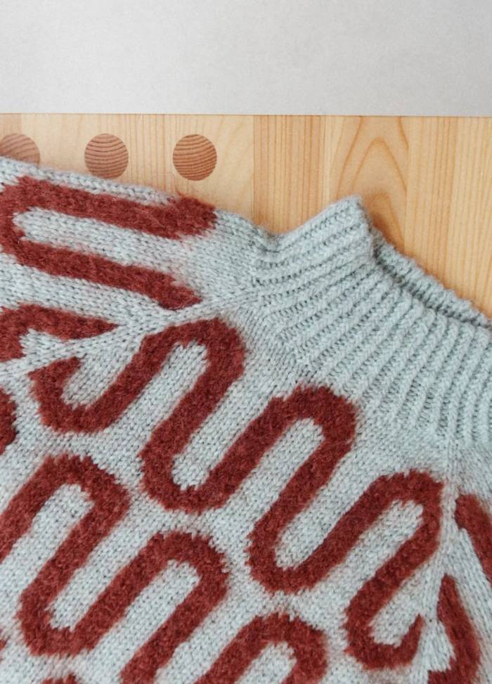 Close-up of a pastel blue knit sweater with a high collar, featuring a red, wavy line pattern. The sweater rests on a wooden surface.