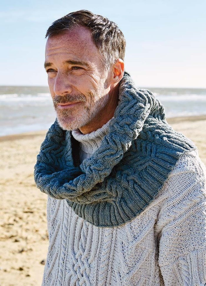 A man with short hair and a beard stands on a beach, wearing a textured gray sweater and scarf. He gazes thoughtfully into the distance under a clear sky.