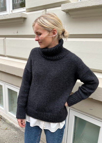 A person in a cozy, dark grey turtleneck sweater stands outside against a beige wall, looking down with a relaxed expression, exuding a calm tone.