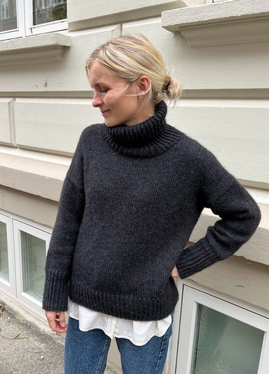 A person in a cozy, dark grey turtleneck sweater stands outside against a beige wall, looking down with a relaxed expression, exuding a calm tone.