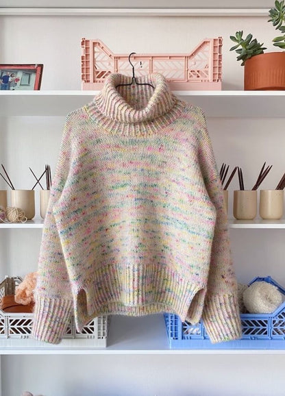 A multicoloured knit sweater with a thick turtleneck and speckled pastel hues hangs on a hanger. It's displayed on white shelves with yarn, knitting needles, and a potted plant, conveying a cozy, handmade vibe.