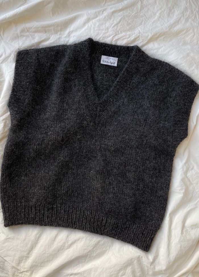 Dark grey, sleeveless V-neck sweater vest on a crumpled white fabric background. The knit texture and casual setting evoke a cozy, relaxed vibe.