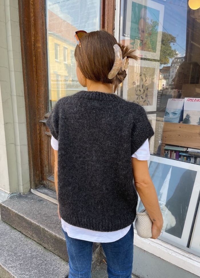 A woman with a hair clip stands outside a storefront, wearing a dark knitted vest over a white shirt and jeans. She holds a woven bag and looks away.