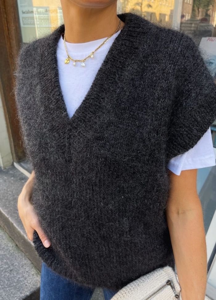 A person wearing a dark grey, fuzzy sleeveless sweater over a white T-shirt stands outdoors. They hold a beige textured bag, exuding a cozy, casual vibe.