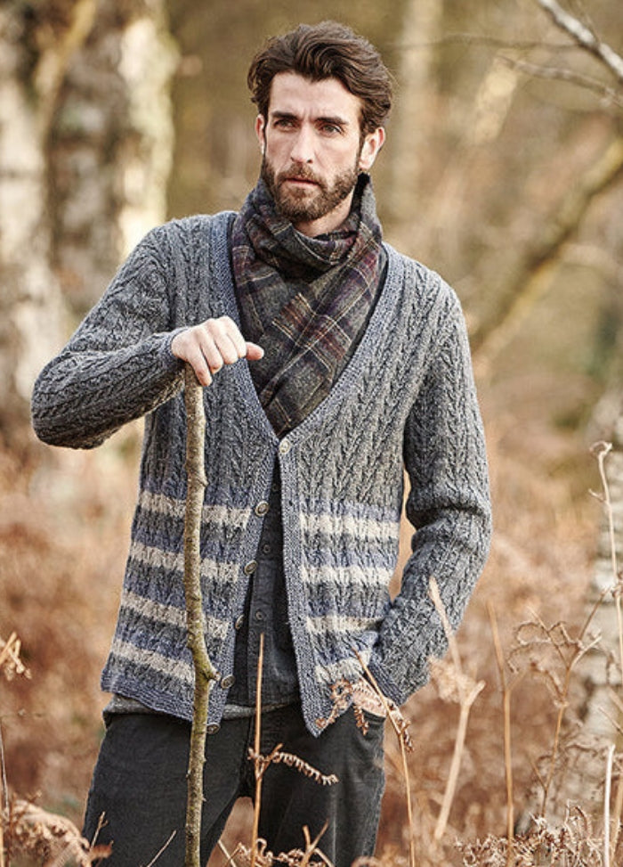 Person wearing the Welara Cardigan, a grey striped knit design, holding a walking stick in a wooded outdoor setting.