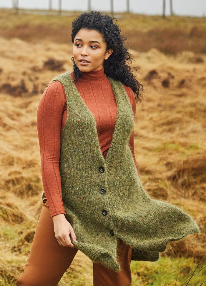 Woman with curly hair wearing an orange sweater and green knitted vest stands in a field of dry grass. She gazes to the side, exuding a calm, contemplative mood.