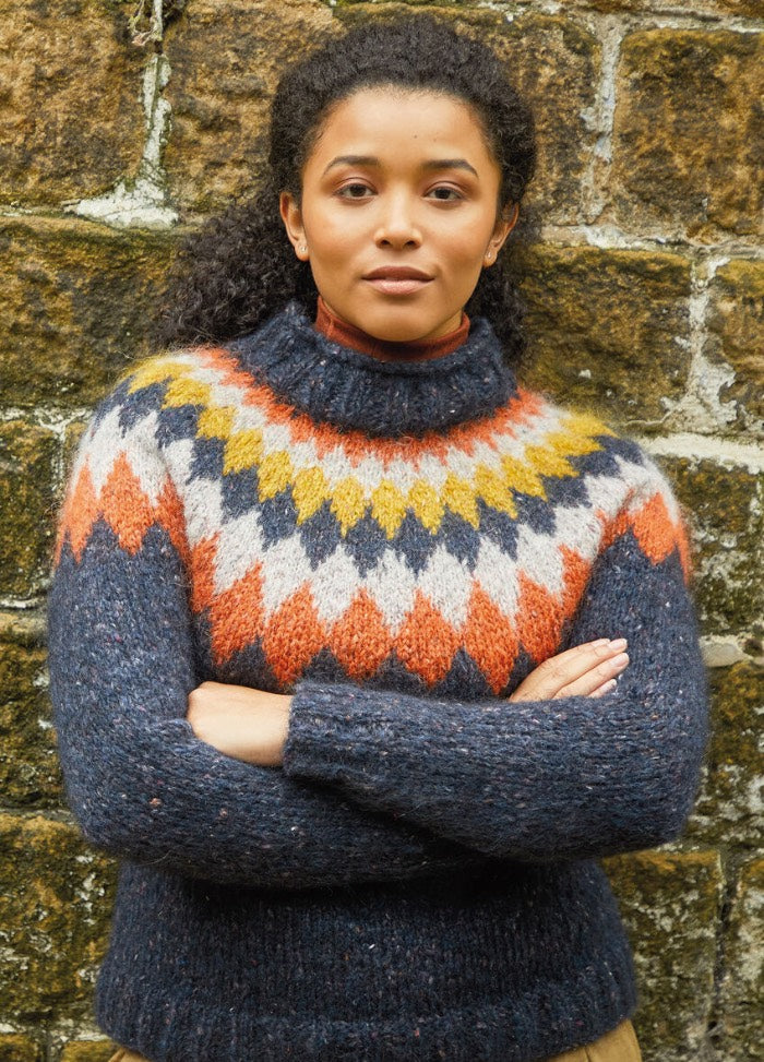 A person in a colorful, patterned sweater stands confidently with arms crossed against a textured stone wall, conveying a calm, self-assured tone.