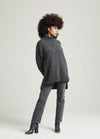 A person with a voluminous afro hairstyle stands confidently in a cozy gray turtleneck sweater, gray jeans, and black ankle boots against a plain backdrop.