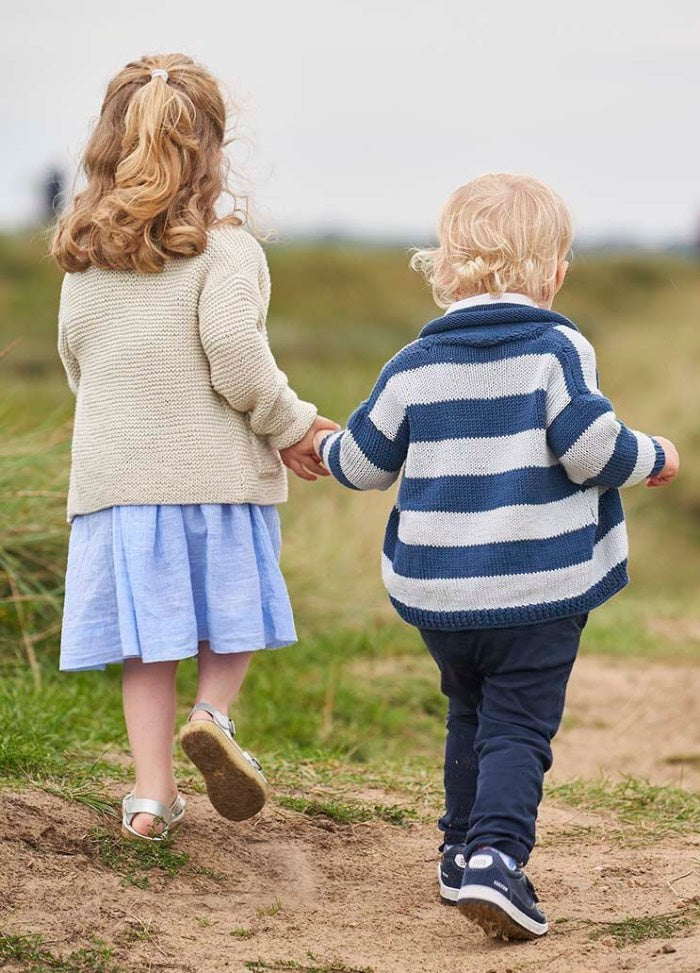 Two children, holding hands, walk on a grassy path. The girl wears a beige sweater and blue dress, while the boy is in a striped sweater and dark pants. Peaceful setting.